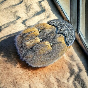 May include: A traditional Scottish sporran, a pouch worn with a kilt. The sporran is made of brown and gray fur with three decorative tassels. A silver-colored metal cantle with Celtic knotwork and a deer design adorns the top.