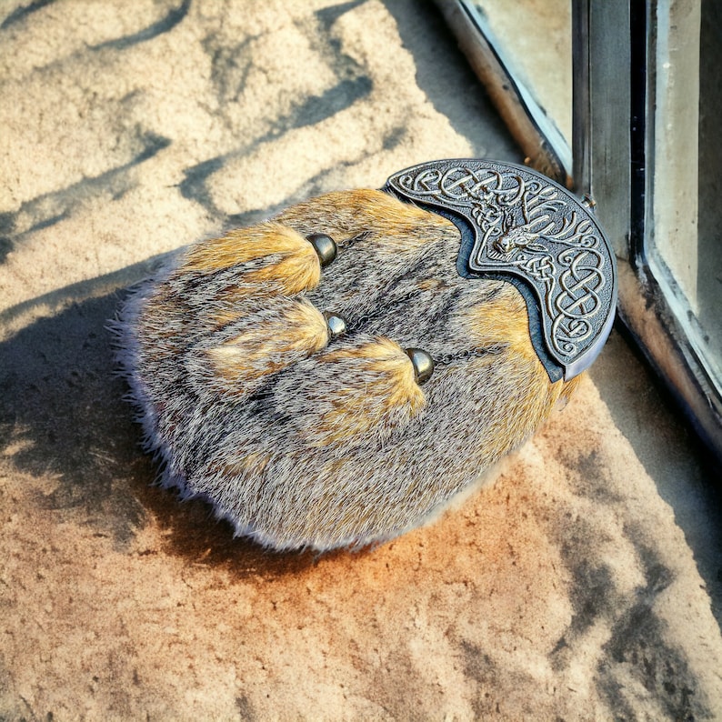 May include: A traditional Scottish sporran, a pouch worn with a kilt. The sporran is made of brown and gray fur with three decorative tassels. A silver-colored metal cantle with Celtic knotwork and a deer design adorns the top.