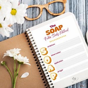 May include: A spiral-bound notebook with the text "The SOAP Bible Study Method" in orange and purple. The notebook is open to a page with lined sections for scripture, observation, application, and prayer. White flowers and wooden glasses are in the background.
