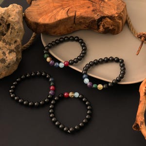 Op de afbeelding: Vier zwarte kralenarmbanden met kleurrijke edelsteenaccenten. De armbanden zijn gerangschikt op een neutraal bord en een donker oppervlak. De edelstenen zijn in een regenboogpatroon gerangschikt.