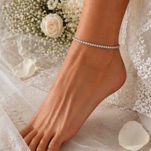 May include: A delicate anklet with small, sparkling stones encircles an ankle. The anklet is set against a backdrop of white flowers and fabric, suggesting a bridal or special occasion theme. The anklet's design is simple yet elegant.