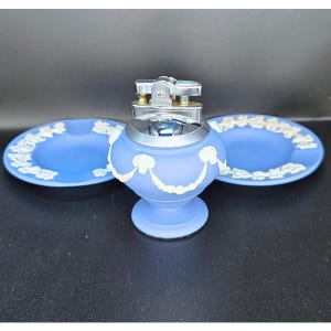 Pode incluir: Um isqueiro azul vintage com uma parte superior prateada e duas cinzeiros a condizer. O isqueiro e os cinzeiros têm um design floral branco. O isqueiro está sobre uma superfície preta.