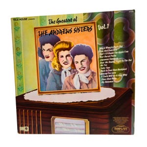 May include: Vintage vinyl record album cover titled "The Greatest of The Andrews Sisters Vol. 1." The cover features a portrait of the singing trio. Song titles are listed on the right side of the cover. The album is displayed on a vintage radio.