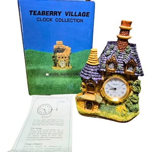 Könnte beinhalten: Eine dekorative Teaberry Village Uhr in Form eines skurrilen Hauses mit lila Dach, goldfarbenem Zifferblatt und Schornstein. Die Uhr steht auf einem grün-gelben Sockel. Auf der Box steht "TEABERRY VILLAGE CLOCK COLLECTION".