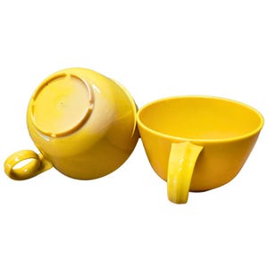May include: Two yellow plastic cups with handles. One cup is upright, the other is upside down. The cups are a bright, solid yellow colour and appear to be made of a durable plastic material. The handles are a simple, curved design.