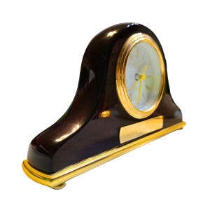 May include: A dark brown, arched mantel clock with a gold-colored bezel and base. The clock face is white with gold hands and numerals. A gold plaque is affixed to the base.
