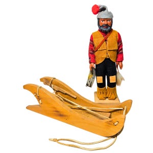 Könnte beinhalten: Ein Holzspielzeugschlitten mit Lederriemen und eine Figur eines bärtigen Mannes in roter und gelber Kleidung, der einen Fisch und eine weiße Feder hält. Der Mann trägt einen grauen Hut mit einem roten Bommel.