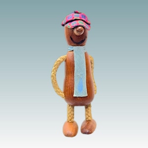 May include: A wooden figurine shaped like a person with a smooth brown body, wearing a light blue scarf and a colorful plaid cap. The figure has braided rope arms and legs, rounded wooden feet, and a painted black smile on its round head.