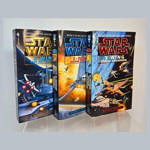 Puede incluir: Tres novelas de Star Wars X-Wing. Los libros presentan portadas vibrantes con escenas de batallas espaciales, el logotipo de Star Wars y los títulos "X-Wing: Rogue Squadron", "X-Wing: Wedge's Gamble" y "X-Wing: The Krytos Trap". El nombre del autor, Michael A. Stackpole, es visible.