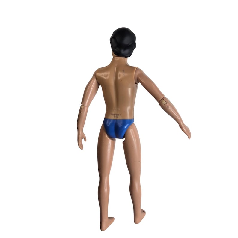 Peut inclure: Une figurine articul&eacute;e en plastique avec un teint bronz&eacute;, des cheveux noirs moul&eacute;s et un maillot de bain bleu. La figurine est vue de dos, pr&eacute;sentant des bras et des jambes articul&eacute;s, avec un corps lisse et des articulations visibles.