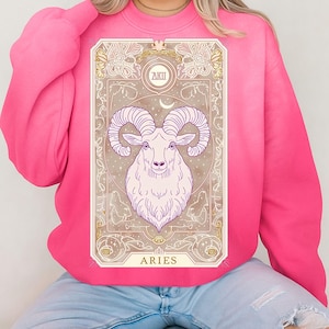 May include: A pink sweatshirt featuring an Aries zodiac design. The design includes a ram illustration within a decorative frame, with the word "ARIES" at the bottom. The sweatshirt is a solid pink color.