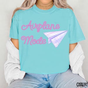 A turquoise t-shirt with the words "Airplane Mode" in pink script and a paper airplane graphic. The shirt is a short-sleeved, casual style.