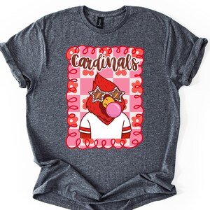 Dark heather gray t-shirt featuring a cartoon cardinal wearing star-shaped sunglasses and blowing a bubblegum bubble. The shirt has the word "Cardinals" in a decorative font within a pink and red floral frame.