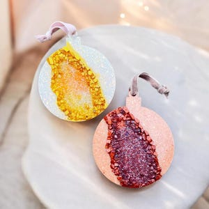 May include: Two round Christmas ornaments with a geode design. One ornament has a white background with a yellow and gold geode design. The other ornament has a pink background with a red and purple geode design. Each ornament has a pink ribbon.