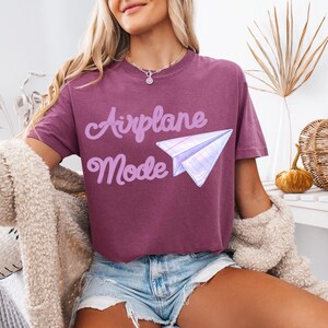A maroon t-shirt with the words "Airplane Mode" in a light purple script and a paper airplane graphic. The shirt is paired with denim shorts and a cream-colored cardigan.