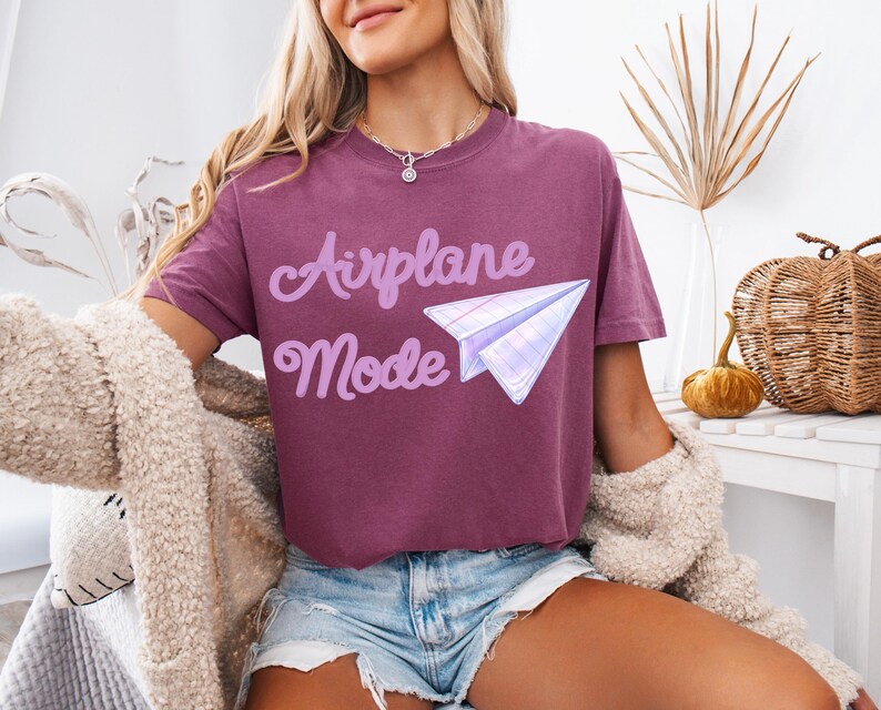 A maroon t-shirt with the words "Airplane Mode" in a light purple script and a paper airplane graphic. The shirt is paired with denim shorts and a cream-colored cardigan.