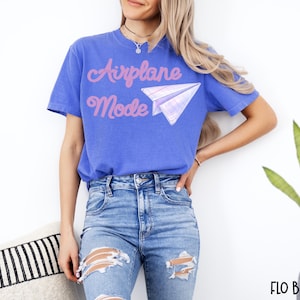 A blue t-shirt with the words "Airplane Mode" in pink script and a paper airplane graphic. The shirt is a solid blue color and is paired with blue jeans.