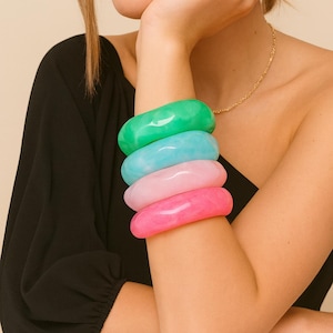 May include: Four colorful bangles in shades of green, blue, pink, and hot pink are stacked on an arm. The bracelets are smooth and glossy, with a rounded rectangular shape. The person is wearing a black top and a gold necklace.