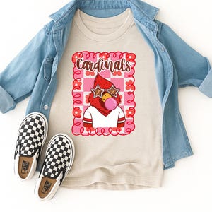 A heather dust colored t-shirt with a graphic of a red cardinal wearing star-shaped sunglasses and blowing a bubblegum bubble. The word "Cardinals" is above the cardinal. A light blue denim jacket and black and white checkered slip-on shoes are also in the image.
