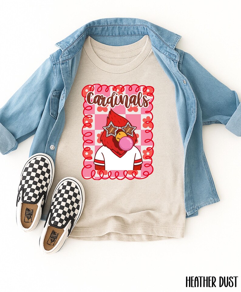 A heather dust colored t-shirt with a graphic of a red cardinal wearing star-shaped sunglasses and blowing a bubblegum bubble. The word "Cardinals" is above the cardinal. A light blue denim jacket and black and white checkered slip-on shoes are also in the image.
