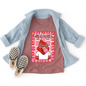heather mauve t-shirt featuring a cartoon cardinal with star-shaped sunglasses blowing a bubble. The cardinal is wearing a white shirt with red stripes. The word "Cardinals" is written in brown script within a pink and red floral frame.