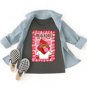 A dark gray t-shirt with a graphic of a red cardinal wearing star-shaped sunglasses and blowing a bubblegum bubble. The shirt is paired with a light blue denim jacket and black and white checkered slip-on shoes. The word "Cardinals" is above the cardinal.