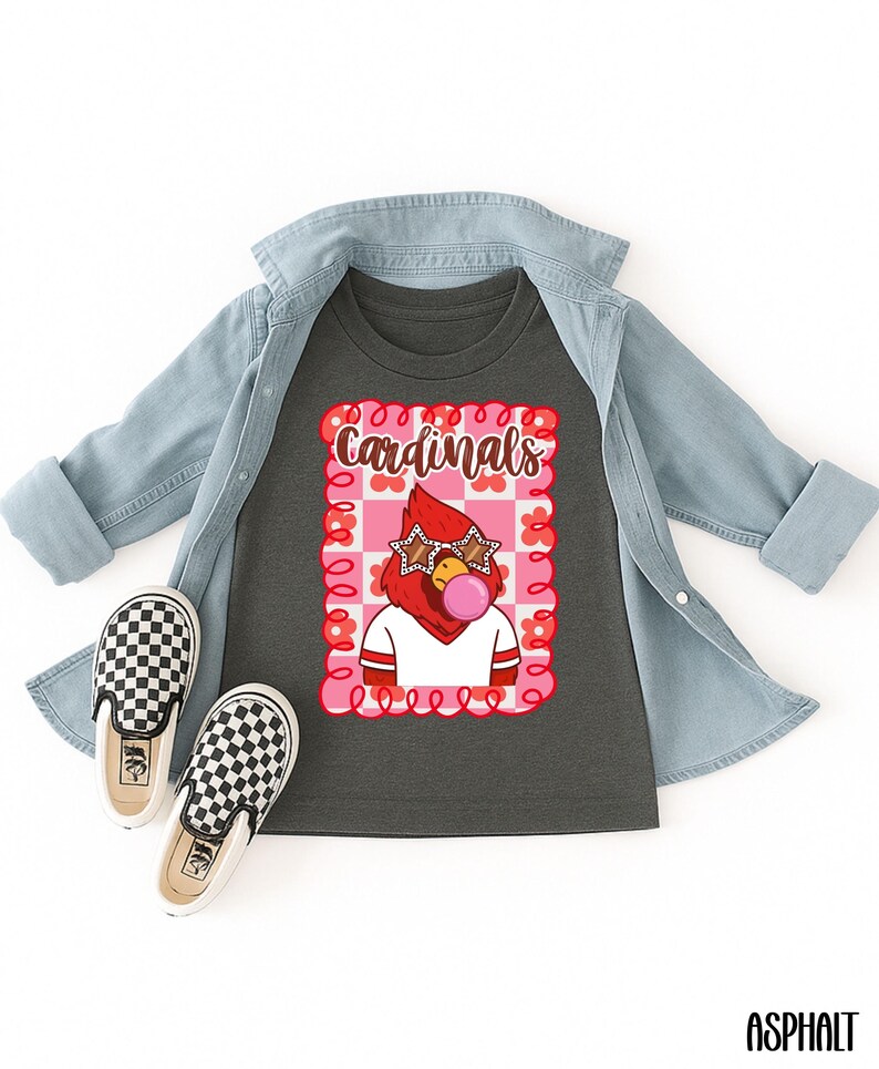 A dark gray t-shirt with a graphic of a red cardinal wearing star-shaped sunglasses and blowing a bubblegum bubble. The shirt is paired with a light blue denim jacket and black and white checkered slip-on shoes. The word "Cardinals" is above the cardinal.