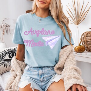 Light blue t-shirt with the text "Airplane Mode" in pink script and a paper airplane graphic. The shirt is made of a soft material and is paired with denim shorts. A beige sweater is draped over the shoulders.