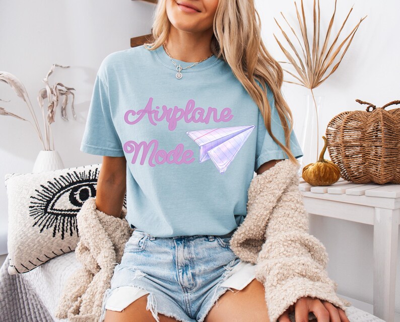 Light blue t-shirt with the text "Airplane Mode" in pink script and a paper airplane graphic. The shirt is made of a soft material and is paired with denim shorts. A beige sweater is draped over the shoulders.