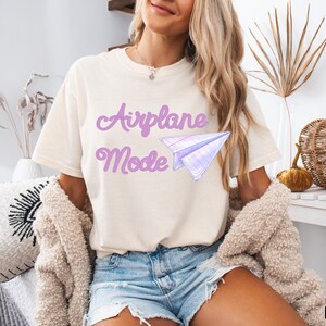 Ivory-colored t-shirt with the words "Airplane Mode" in lavender script and a matching paper airplane graphic. The shirt is a casual style, perfect for everyday wear. The shirt is made of a soft material.