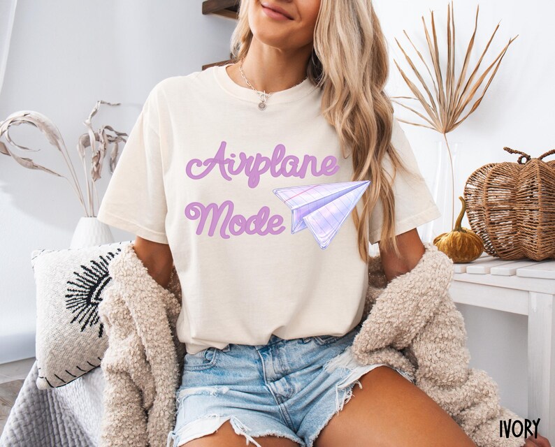 Ivory-colored t-shirt with the words "Airplane Mode" in lavender script and a matching paper airplane graphic. The shirt is a casual style, perfect for everyday wear. The shirt is made of a soft material.