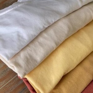 A stack of folded t-shirts in various colors: white, cream, yellow, gold, and rust. The shirts are neatly arranged, showcasing the different hues and textures of the fabric. The image highlights the variety of colors available.