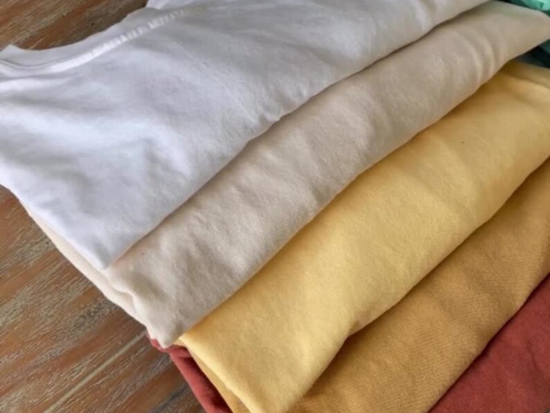 A stack of folded t-shirts in various colors: white, cream, yellow, gold, and rust. The shirts are neatly arranged, showcasing the different hues and textures of the fabric. The image highlights the variety of colors available.