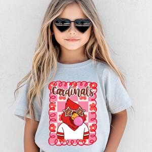 A light gray t-shirt featuring a cartoon cardinal wearing star-shaped sunglasses and blowing a bubblegum bubble. The word "Cardinals" is written above the graphic in a decorative font, framed by a pink and red floral design.