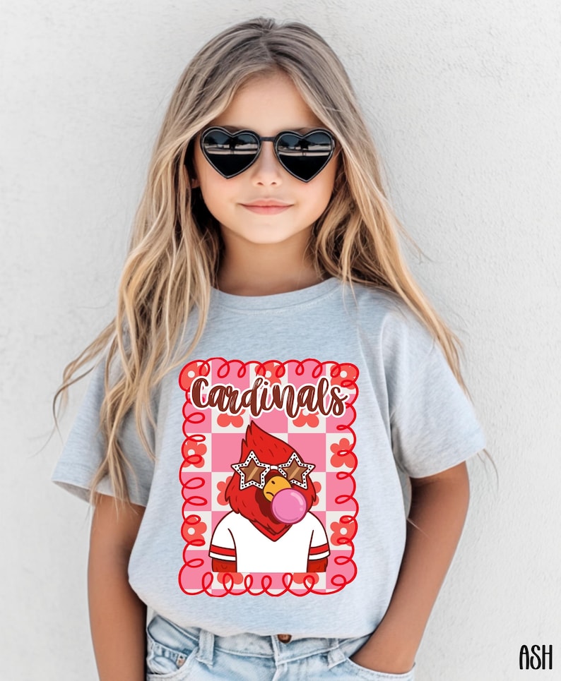 A light gray t-shirt featuring a cartoon cardinal wearing star-shaped sunglasses and blowing a bubblegum bubble. The word "Cardinals" is written above the graphic in a decorative font, framed by a pink and red floral design.