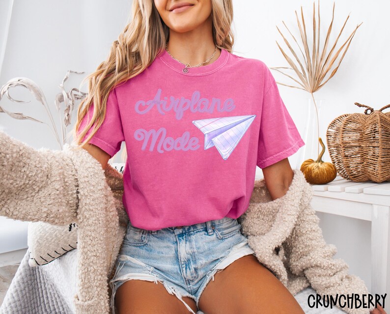 May include: A magenta t-shirt with the words "Airplane Mode" and a paper airplane graphic in light purple. The shirt is paired with denim shorts and a cream-colored cardigan. The word "CRUNCHBERRY" is in the bottom right corner.