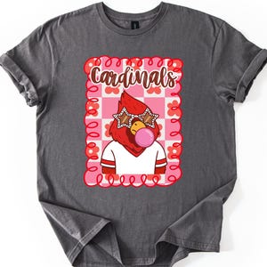 Charcoal-colored t-shirt featuring a cartoon cardinal with star-shaped sunglasses blowing a bubblegum bubble. The design includes the word "Cardinals" in a playful font, set within a pink and red floral frame. The shirt is a comfortable fit.