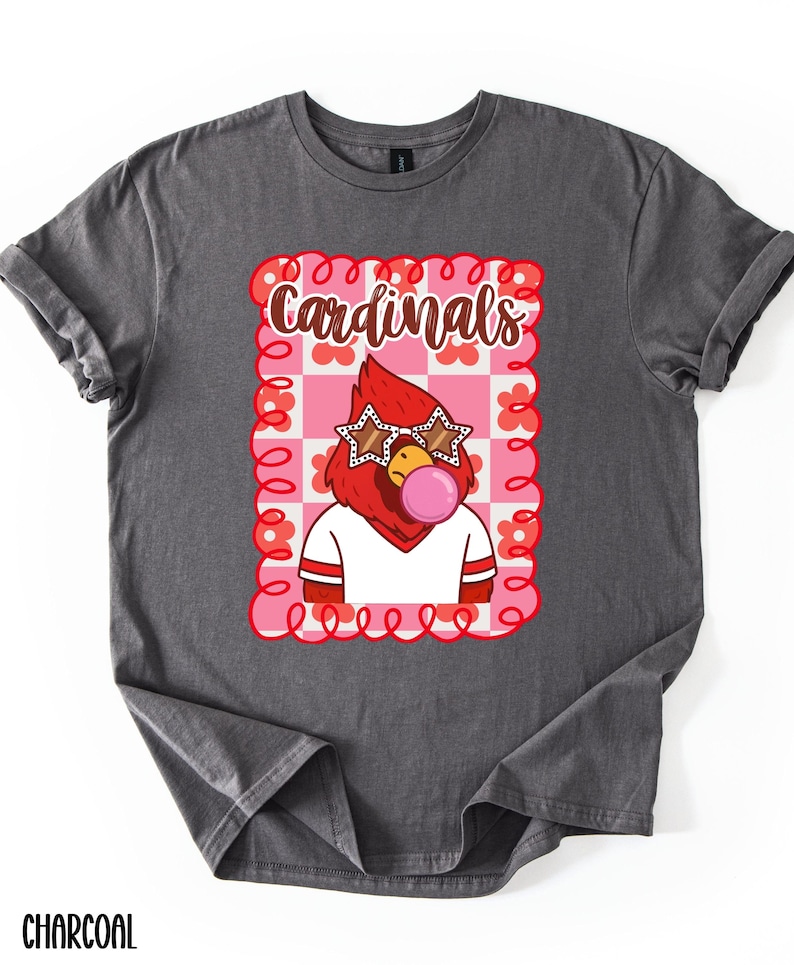 Charcoal-colored t-shirt featuring a cartoon cardinal with star-shaped sunglasses blowing a bubblegum bubble. The design includes the word "Cardinals" in a playful font, set within a pink and red floral frame. The shirt is a comfortable fit.