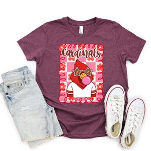 Heather maroon t-shirt with a graphic of a cardinal wearing star-shaped sunglasses and blowing a bubble. The word "Cardinals" is above the graphic. White sneakers and denim shorts are next to the shirt.