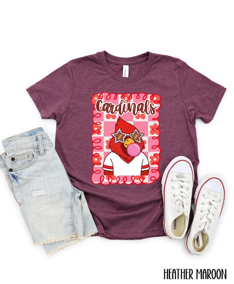 Heather maroon t-shirt with a graphic of a cardinal wearing star-shaped sunglasses and blowing a bubble. The word "Cardinals" is above the graphic. White sneakers and denim shorts are next to the shirt.
