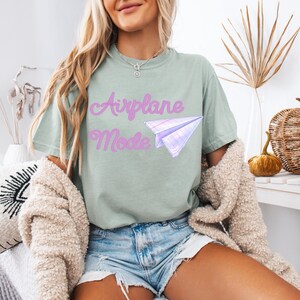 Sage green t-shirt with the text "Airplane Mode" in a cursive font and a paper airplane graphic. The shirt is paired with denim shorts and a cream-colored cardigan.