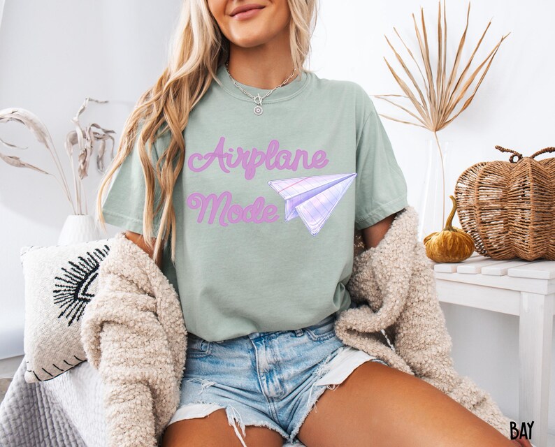 Sage green t-shirt with the text "Airplane Mode" in a cursive font and a paper airplane graphic. The shirt is paired with denim shorts and a cream-colored cardigan.
