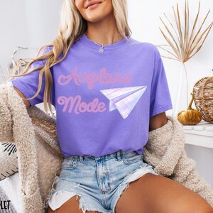 Violet-colored t-shirt with the words "Airplane Mode" in pink script and a paper airplane graphic. The shirt is a short-sleeved, casual style.