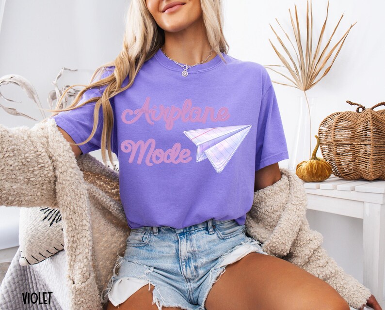 Violet-colored t-shirt with the words "Airplane Mode" in pink script and a paper airplane graphic. The shirt is a short-sleeved, casual style.