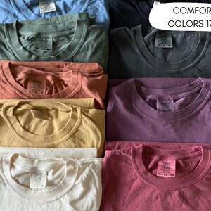 A collection of folded t-shirts in various colors, including blue, green, orange, yellow, white, purple, and pink. The shirts are arranged in a neat stack, with the text "COMFORT COLORS 1717" visible in the upper right corner.