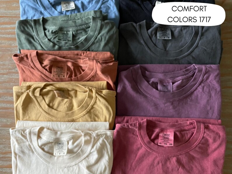 A collection of folded t-shirts in various colors, including blue, green, orange, yellow, white, purple, and pink. The shirts are arranged in a neat stack, with the text "COMFORT COLORS 1717" visible in the upper right corner.