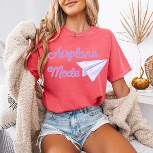 A coral-colored t-shirt with the words "Airplane Mode" in purple script and a paper airplane graphic. The shirt is short-sleeved and has a relaxed fit. The model is wearing denim shorts and a cream-colored cardigan.