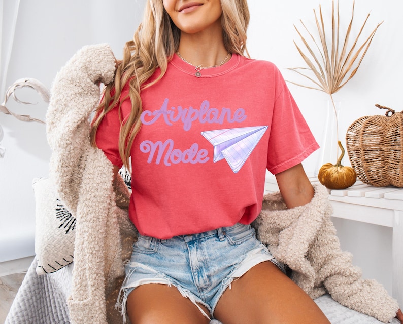 A coral-colored t-shirt with the words "Airplane Mode" in purple script and a paper airplane graphic. The shirt is short-sleeved and has a relaxed fit. The model is wearing denim shorts and a cream-colored cardigan.
