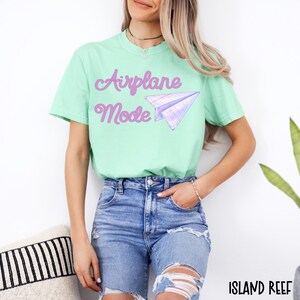 A light green t-shirt with the words "Airplane Mode" in purple script and a purple paper airplane graphic. The model is wearing blue jeans with ripped knees.