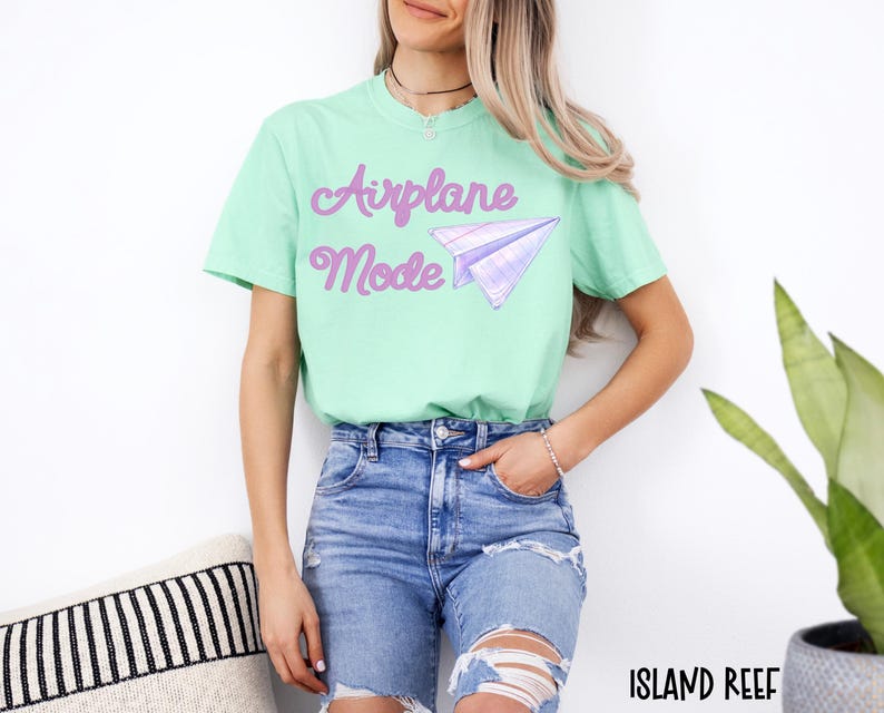A light green t-shirt with the words "Airplane Mode" in purple script and a purple paper airplane graphic. The model is wearing blue jeans with ripped knees.